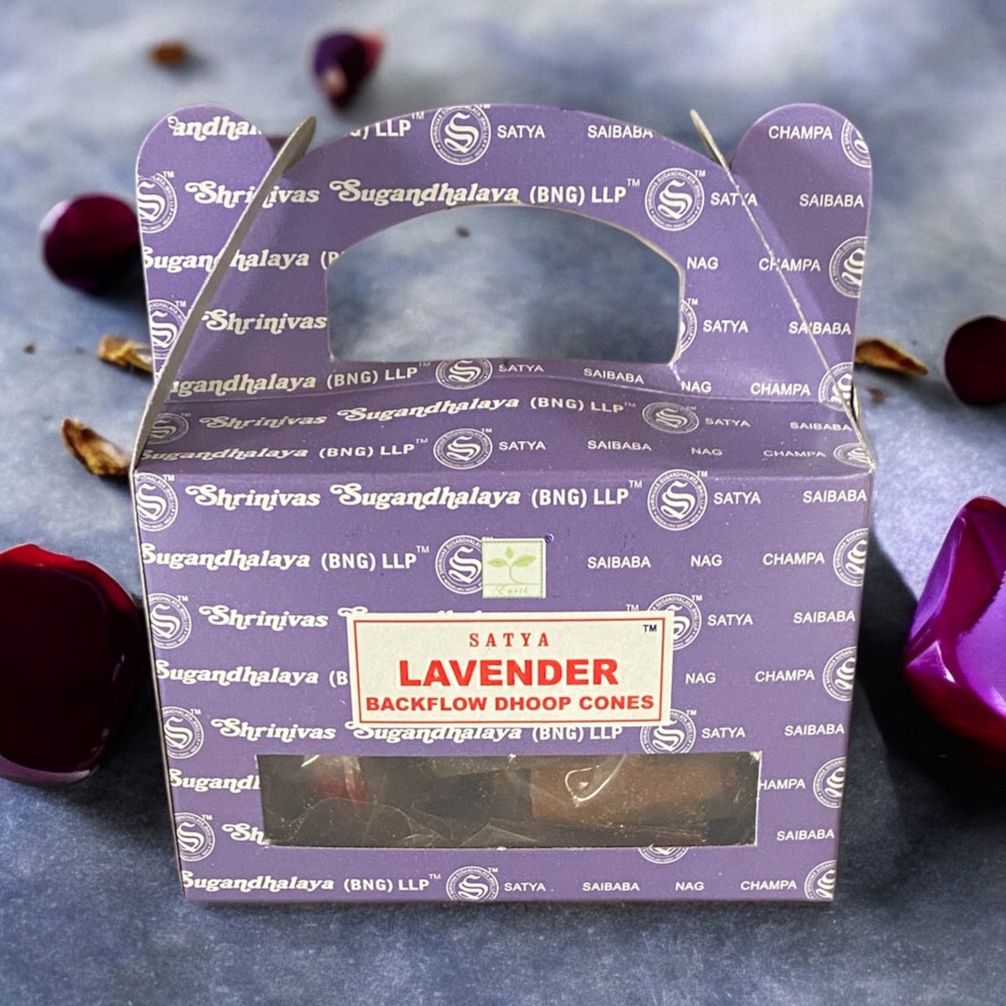A box of Satya Lavender Backflow Dhoop Cones with purple packaging, displayed on a blue background with scattered rose petals around.