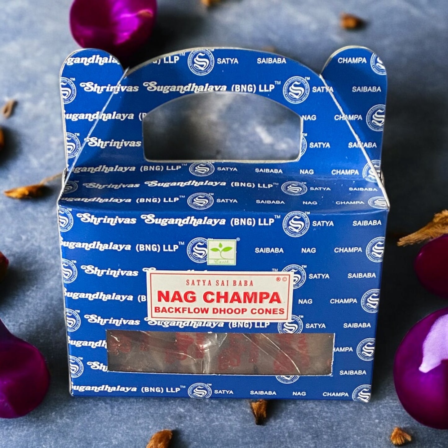 A box of Nag Champa Backflow Dhoop Cones with a blue label, displayed on a surface with purple flowers scattered around it.