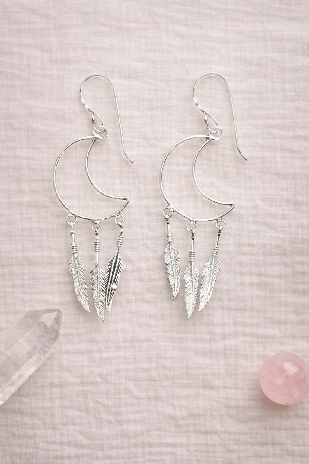 Silver moon-shaped dream catcher earrings with feather details on a light pink background