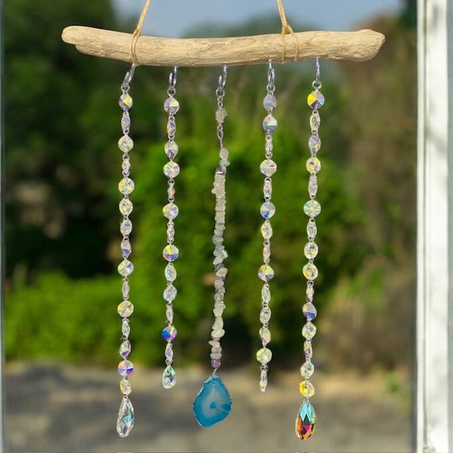 A handmade driftwood suncatcher with hanging crystal droppers and a central agate slice, displayed against an outdoor backdrop.