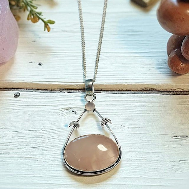 Teardrop-shaped pendant with a pink stone on a silver chain, placed on a textured surface.