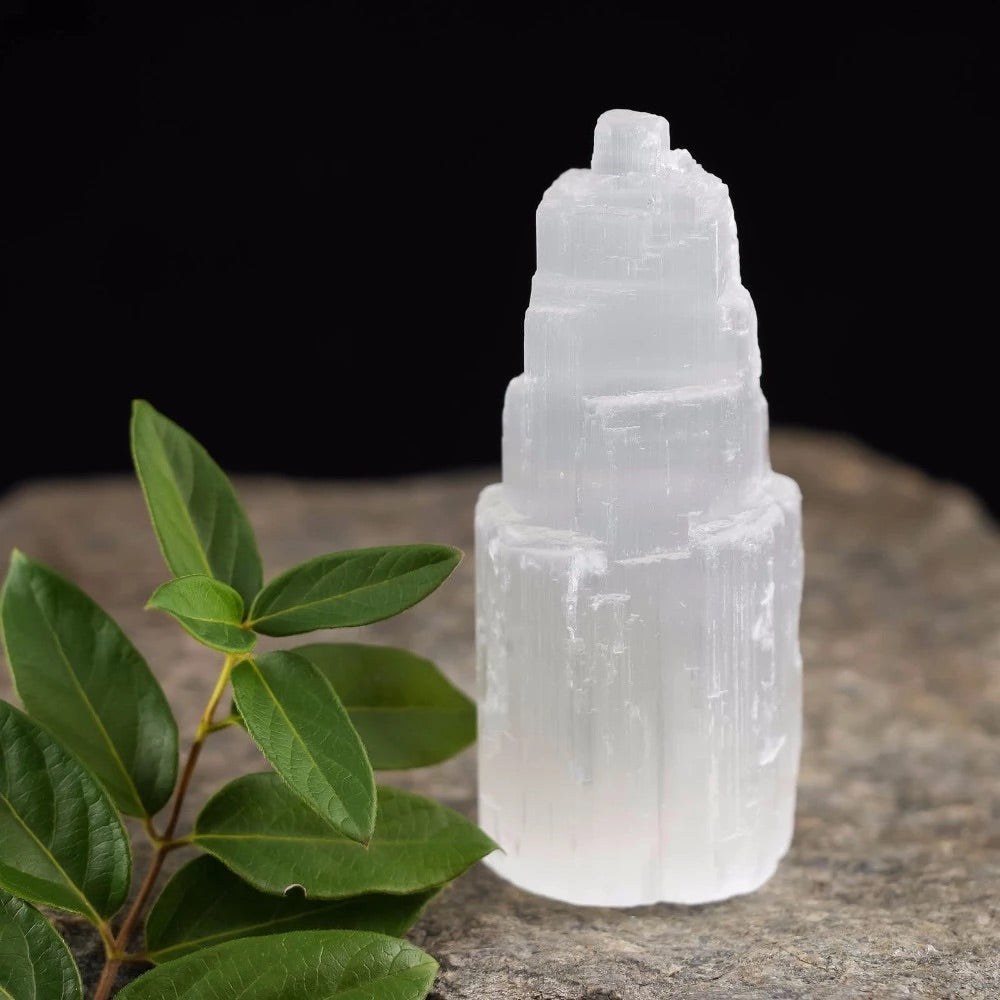 A clear selenite crystal tower placed on a rock, with green leaves in the background.