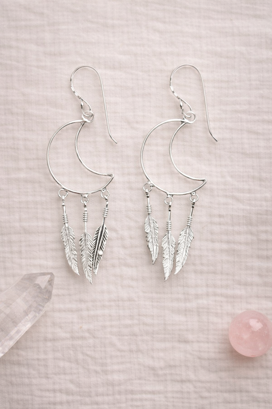Silver moon-shaped dream catcher earrings with feather details on a light pink background