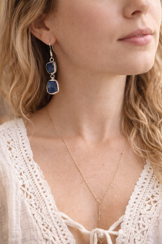 Close-up of a woman wearing blue  Kyanite earrings and a delicate necklace, with a blurred background.