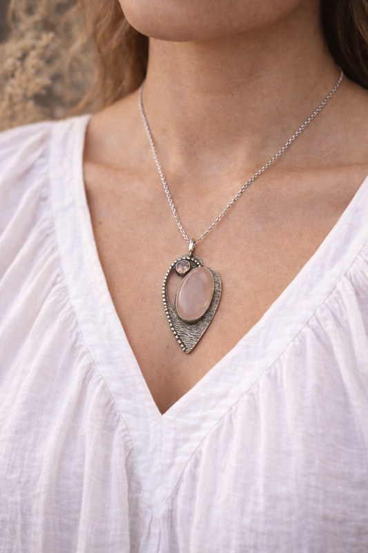 Heart shaped Necklace with a  rose quartz inset pendant worn by a person in a white top, against a blurred natural background.