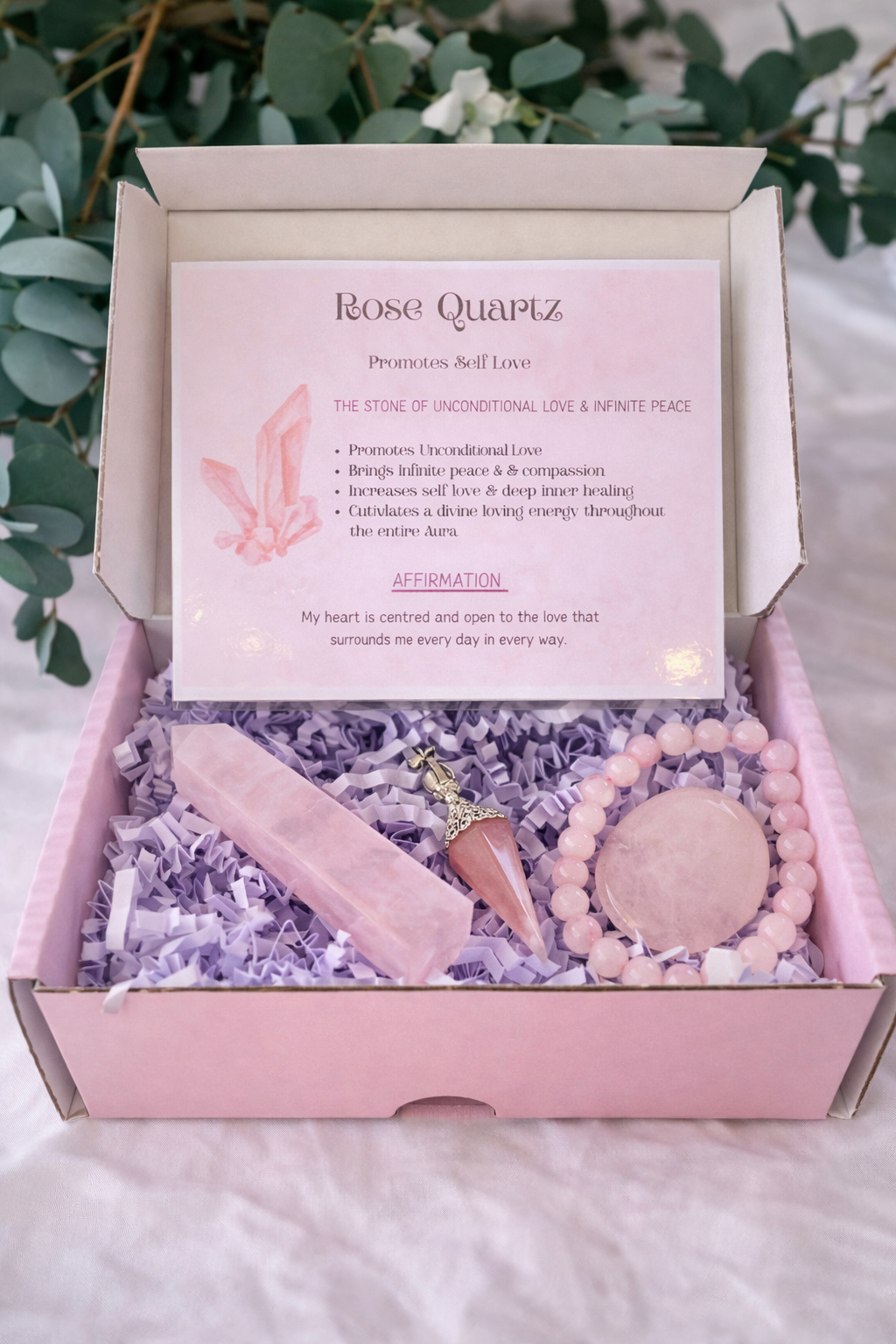 Pink 'Unconditional Love Box' with crystals and a bracelet on a white fabric background with greenery.