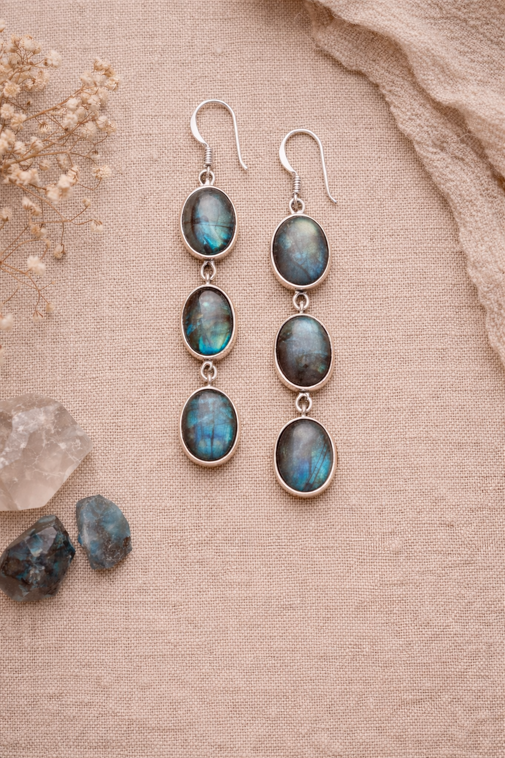 Silver earrings with Labradorite stones on a textured beige surface with dried flowers and crystals.