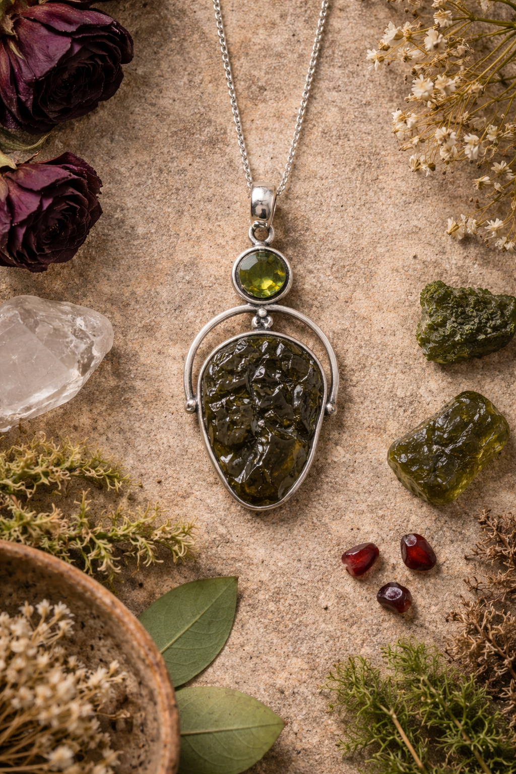 Silver pendant with green Moldavite gemstone on a textured surface with dried flowers and crystals