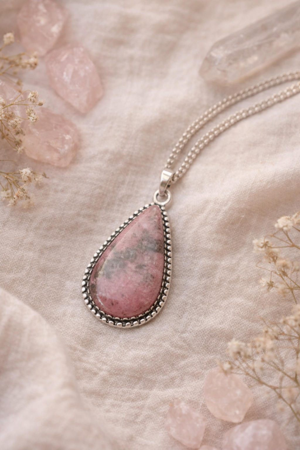 Necklace with a pink teardrop rhodonite pendant on a soft, light-colored surface with dried flowers.