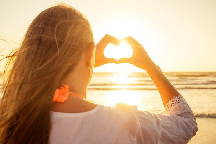 Sunset beach woman making heart hands — self-love, peace, and light.