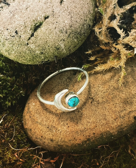 A sterling silver moon ring with a turquoise gemstone, placed on a rocky surface with natural surroundings.
