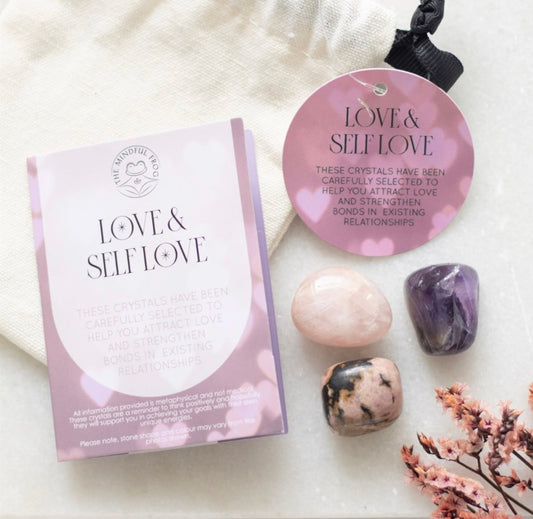 A set of pink-colored healing crystals displayed alongside a pink packaging that reads 'LOVE & SELF-LOVE'. The crystals include a rose quartz, an amethyst, and a rhodonite.
