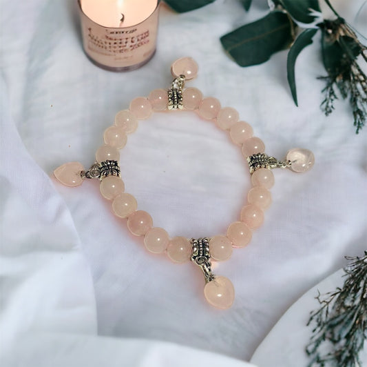 A  rose quartz bracelet with four heart-shaped rose quartz beads and silver accents, displayed on a white background with a candle and greenery in the background.