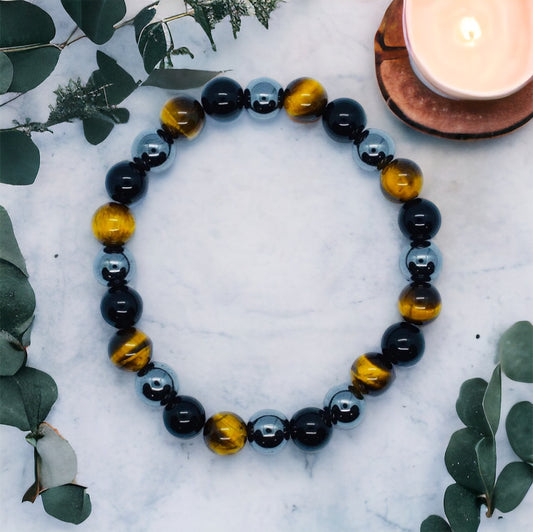 A multicolored bead bracelet featuring a mix of tiger's eye, black obsidian, and hematite beads, displayed on a white surface with greenery in the background and a candle in the foreground.