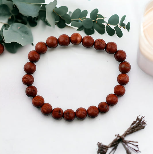 A red jasper bead bracelet displayed on a white surface with green leaves in the background.
