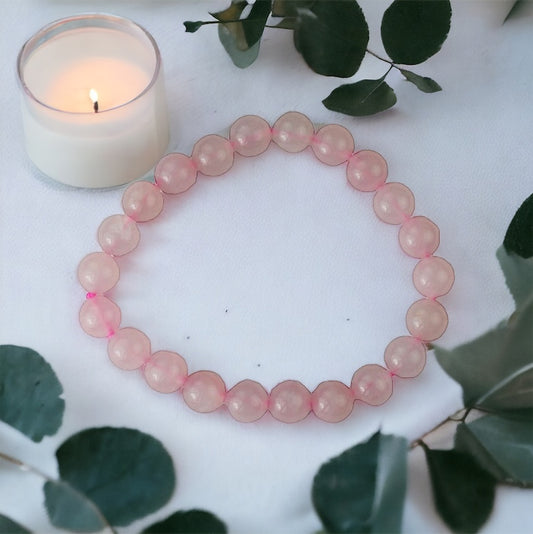 A rose quartz power bead bracelet displayed on a surface with a candle and green leaves in the background, signifying its use for healing and spiritual purposes.