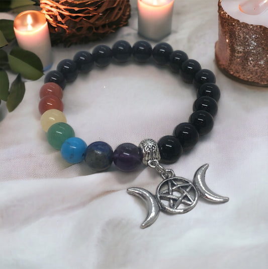 A bracelet with multiple chakra crystal beads and a triple moon cuff charm, presented against a backdrop with a candle and decorative elements.